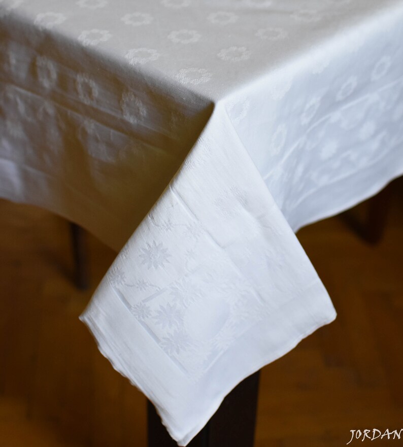 May include: White damask tablecloth with a floral pattern. The tablecloth is folded over a table and the corner is hanging down.