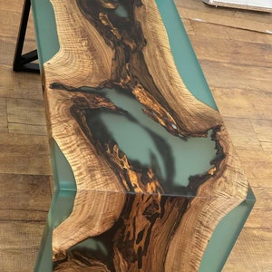 May include: A wooden coffee table with a natural wood grain finish and a large, clear epoxy resin river running through the centre. The resin is a light blue colour and has a smooth, glassy finish.