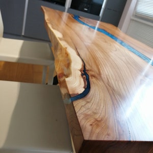 May include: A close-up of a wooden table with a blue epoxy resin river running through the center. The table has a live edge and a natural wood finish.
