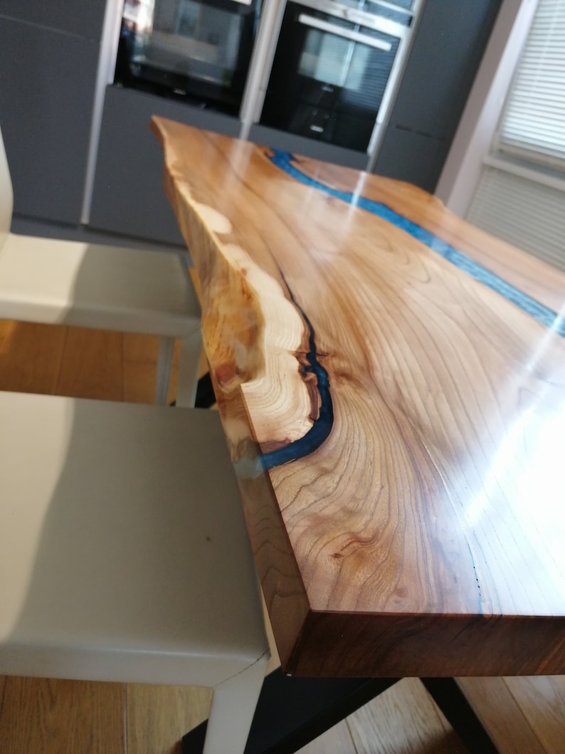 May include: A close-up of a wooden table with a blue epoxy resin river running through the center. The table has a live edge and a natural wood finish.