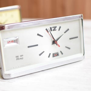May include: A rectangular, silver-framed clock with a white face. The clock features black hour markers, black hands, and a red second hand. The date is displayed as "24" in a small window. The text "DG ST Hero" is printed on the face.