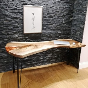 Pu&ograve; includere: Una scrivania con bordo vivo con un piano in legno naturale e un'intarsio in resina epossidica trasparente. La scrivania ha gambe a forcina nere e si trova su un pavimento in legno. Un computer portatile &egrave; sulla scrivania.