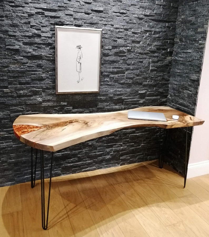 Pu&ograve; includere: Una scrivania con bordo vivo con un piano in legno naturale e un'intarsio in resina epossidica trasparente. La scrivania ha gambe a forcina nere e si trova su un pavimento in legno. Un computer portatile &egrave; sulla scrivania.