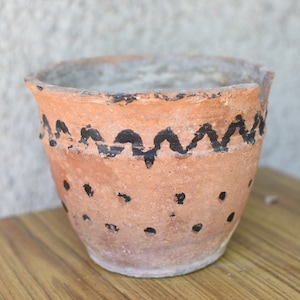 Puede incluir: Una maceta de terracota con un diseño ondulado negro alrededor de la parte superior y puntos negros dispersos por los lados.