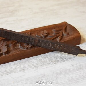 May include: A metal rasp with a wooden handle rests on a carved wooden base. The rasp is dark brown with a textured surface. The base is also brown and features floral carvings. The word "JORDAN" is visible.