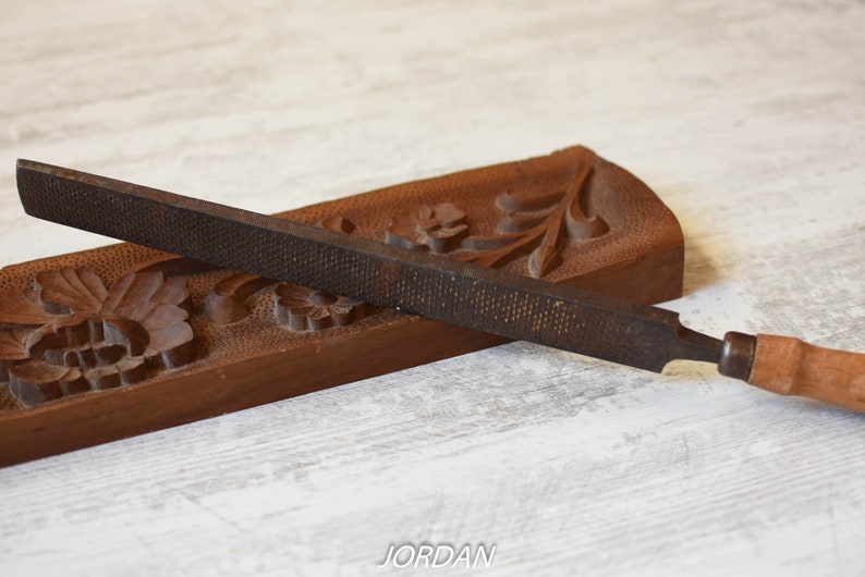 May include: A metal rasp with a wooden handle rests on a carved wooden base. The rasp is dark brown with a textured surface. The base is also brown and features floral carvings. The word "JORDAN" is visible.