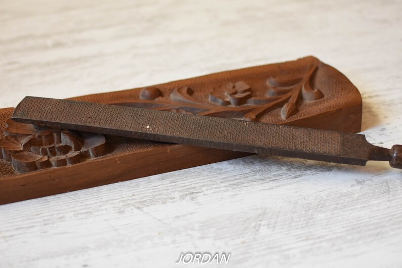 May include: A vintage woodworking rasp and a carved wooden tray. The rasp is a dark brown color with a textured surface. The tray is also brown and features carved floral and leaf designs. The rasp is approximately 10 inches long.