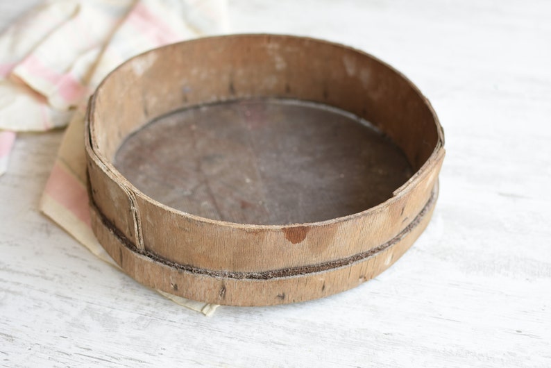 May include: A round wooden sifter with a worn, rustic appearance. The sifter is made of natural wood and has a smooth, slightly textured surface.