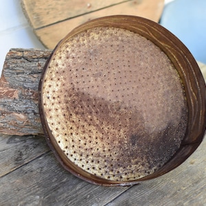 Puede incluir: Un regadera de metal oxidado con una parte superior redonda y perforada. La regadera est&aacute; sobre una superficie de madera.