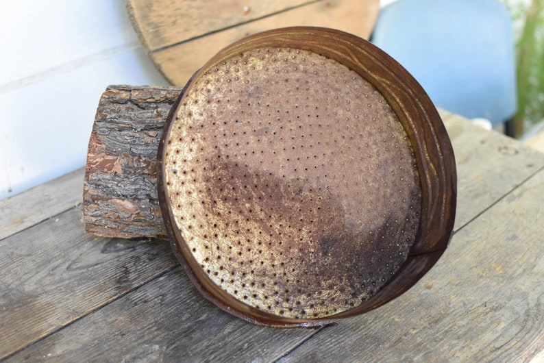 Puede incluir: Un regadera de metal oxidado con una parte superior redonda y perforada. La regadera est&aacute; sobre una superficie de madera.