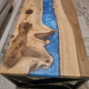 May include: A wooden coffee table with a blue resin river running down the center. The table has a black metal geometric base.