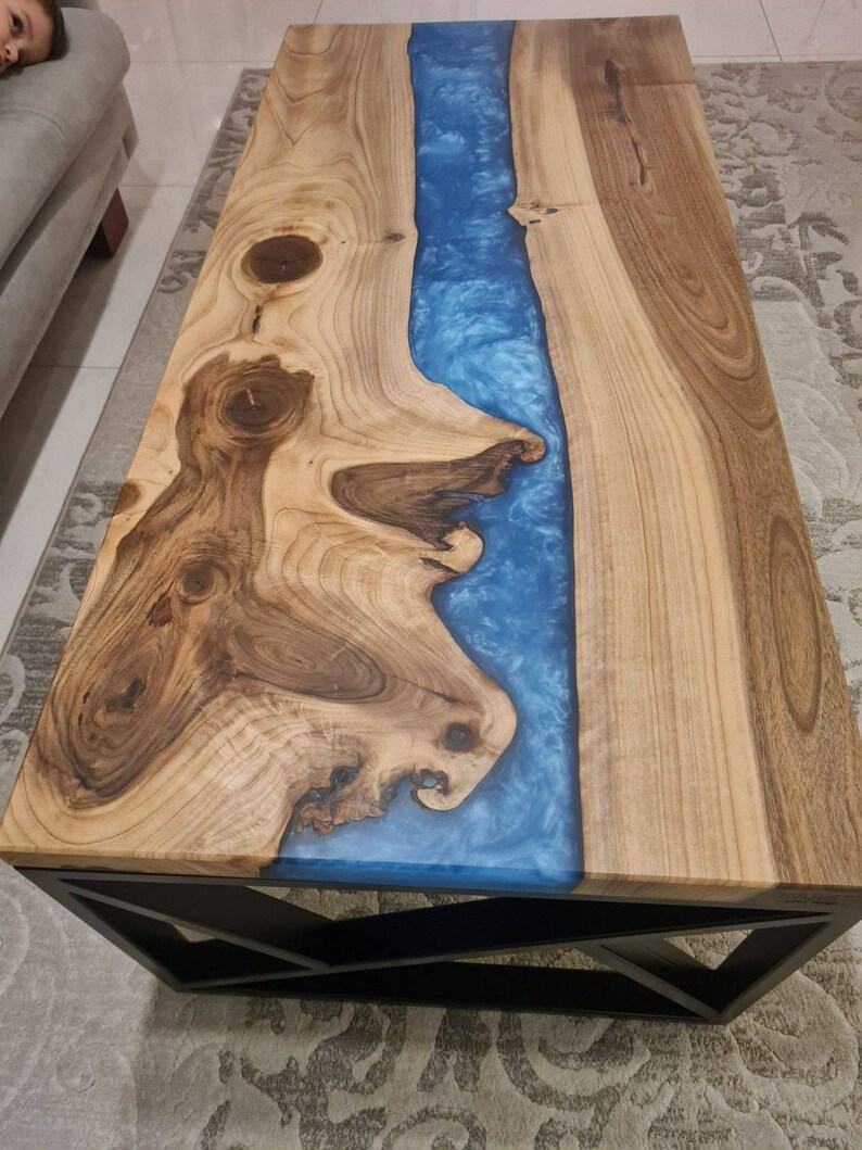 May include: A wooden coffee table with a blue resin river running down the center. The table has a black metal geometric base.