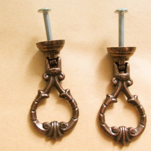 Puede incluir: Dos pomos de armario de bronce ornamentados con un diseño decorativo en forma de lágrima. Cada pomo tiene un tornillo para sujetarlo a una puerta de armario.