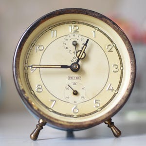 May include: Vintage Peter alarm clock with a cream-colored face and black hands. The clock has a brass-colored bezel and feet, with the word "PETER" printed on the face. The clock is set to approximately 8:45.