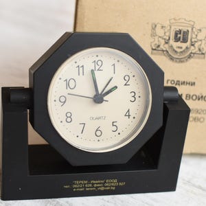 May include: A black octagonal clock with a white face and black hands. The clock is on a black stand with a gold inscription that reads "TEREM - Ivailo EOOD tel.: 062/21 826, fax: 062/1623 527 e-mail terem_vida@mail.bg".