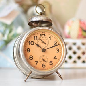 May include: A vintage silver alarm clock with a peach-colored face. The clock has black hands and numbers, and a small seconds dial. It stands on three small legs and has a bell on top. The clock is a classic design.