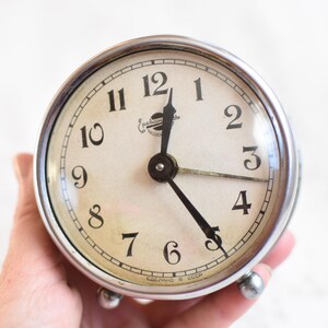May include: A silver alarm clock with black hands and black numbers on a white face. The clock has a chrome finish and a glass face. The clock is set to 3:55.