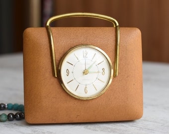 May include: A vintage brown leather clock with a gold metal frame and a white face. The clock has a gold handle and is set on a white surface.