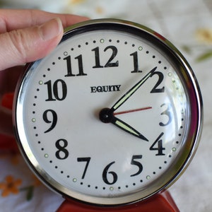 May include: A silver alarm clock with a white face and black numbers. The clock is set to 1:55 and has the word "EQUITY" printed on the face.