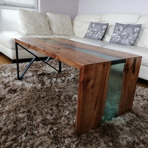 May include: A wooden coffee table with a clear epoxy river running through the center. The table has a black metal frame and is sitting on a brown shag rug.