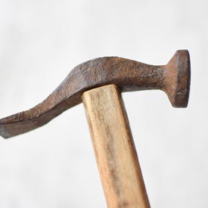 May include: A vintage hammer featuring a weathered, rusted iron head with a flat striking face and a curved claw. The head is securely attached to a light-colored, smooth wooden handle, showing signs of age and consistent use over time.