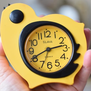 May include: A yellow and black vintage alarm clock in the shape of a bird. The clock face is yellow with black numerals and hands, and the brand name "SLAVA" is printed on the face. The clock is held in a hand.
