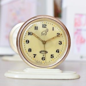 Può includere: Un orologio da tavolo vintage bianco con un bordo dorato e un logo di elefante sul quadrante. Il quadrante dell'orologio ha numeri e lancette neri, e il testo "OPYHKBA" è stampato sotto i numeri.