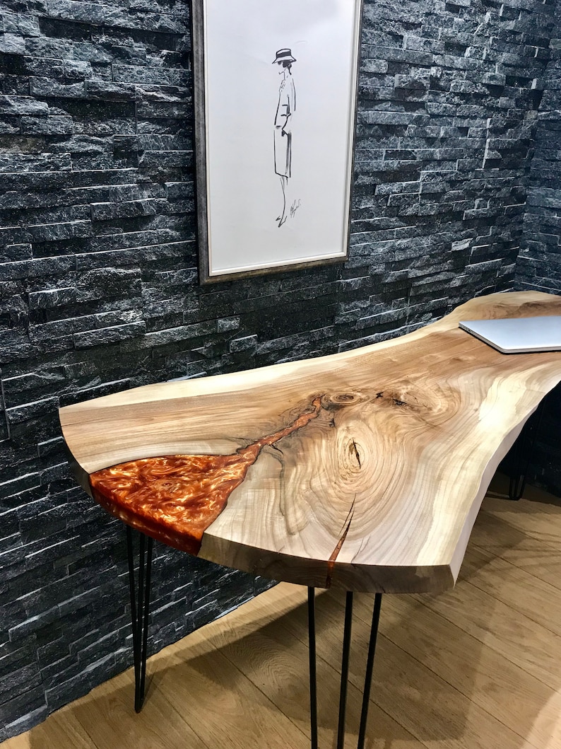 Pu&ograve; includere: Una scrivania in legno a bordo vivo con un inserto in resina. La scrivania ha una finitura in legno naturale ed &egrave; sostenuta da gambe a forcina nere.