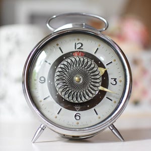 May include: A round silver-toned alarm clock with a metallic frame and two small feet. The clock face features a white background, black numerals, and a central circular design with a black and white spiral pattern. Gold hands indicate the time.