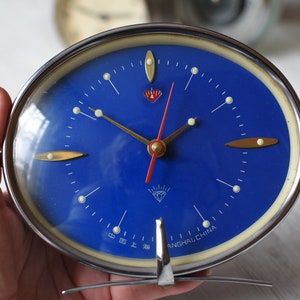 May include: A vintage blue alarm clock with a white face and gold hands. The clock has a diamond logo and the text "SHANGHAI, CHINA" on the face.
