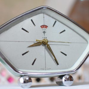 May include: A pentagonal table clock with a chrome frame and two spherical chrome feet. The off-white face features black hour markers, gold-toned hands, and a red logo at the top with the text Polaris and Chinese characters below.