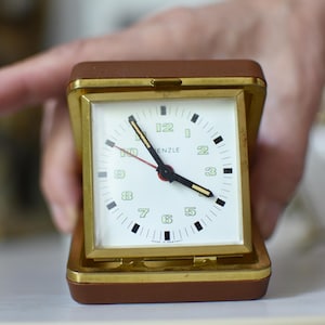 May include: A square travel alarm clock with a brown leather-like case and a gold-tone metal frame. The white clock face features green Arabic numerals, black hour and minute hands with yellow tips, a red second hand, and the text "Kienzle".