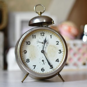 May include: A round silver-toned analog alarm clock with a white face, black Arabic numerals, and a top bell mechanism. The clock features thin black hands, a small sub-dial at the bottom, and two slender brass-colored legs for support.