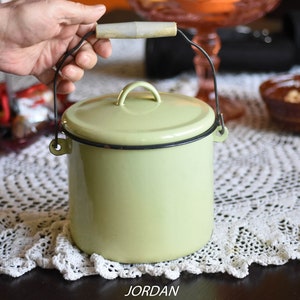 Könnte beinhalten: Ein hellgrüner Emaillebehälter mit schwarzem Rand und einem Holzgriff. Der Behälter hat einen Deckel und einen schwarzen Drahtgriff.