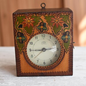 May include: A wooden clock with a painted floral design. The clock face is white with black numbers and hands. The clock is approximately 12.5 cm tall.