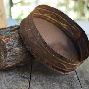 Puede incluir: Una bandeja redonda de metal oxidado con un borde decorativo. La bandeja est&aacute; sentada sobre un trozo de madera.