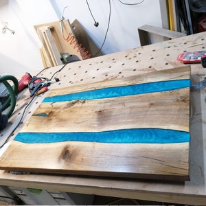 Puede incluir: Una encimera de mesa de madera con dos incrustaciones de r&iacute;o de resina azul. La encimera est&aacute; sobre un banco de trabajo en un taller.