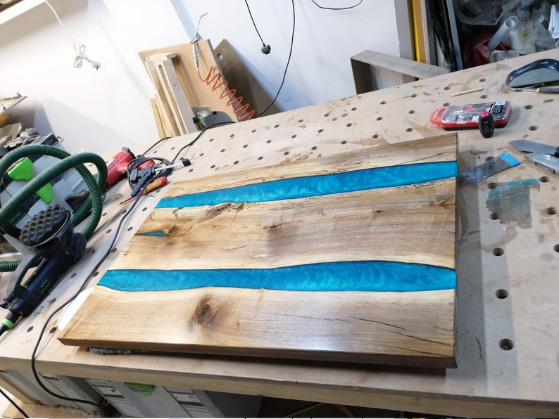 Puede incluir: Una encimera de mesa de madera con dos incrustaciones de r&iacute;o de resina azul. La encimera est&aacute; sobre un banco de trabajo en un taller.