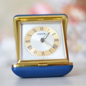 May include: A gold-toned, square travel clock with a white face and Roman numerals. The clock face has the word "EQUITY" printed on it. The clock is encased in a blue and gold frame.