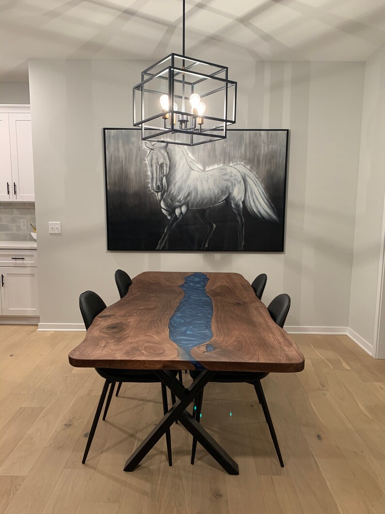 May include: A dark wood dining table with a blue resin river inlay and black metal X-shaped legs. The table is set with four black chairs. A black caged light fixture hangs above the table. A black and white painting of a horse hangs on the wall behind the table.