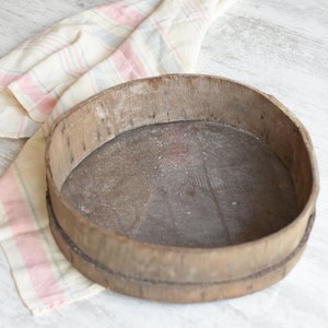 May include: A round wooden sifter with a woven mesh bottom. The sifter is made of natural wood and has a rustic appearance. It is sitting on a white surface with a pink and white striped cloth.