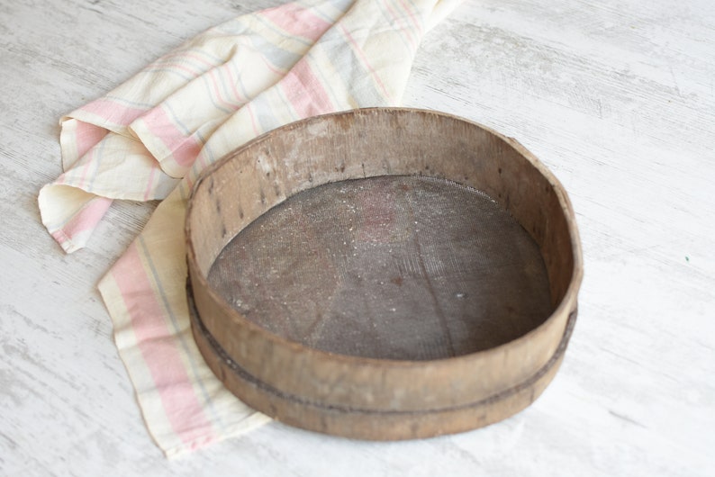 May include: A round wooden sifter with a woven mesh bottom. The sifter is made of natural wood and has a rustic appearance. It is sitting on a white surface with a pink and white striped cloth.