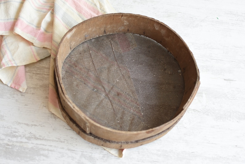 May include: A round wooden sifter with a mesh screen. The sifter is made of natural wood and has a rustic, vintage look.