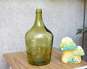 Puede incluir: Una gran botella de vidrio verde con una boca ancha se encuentra sobre una mesa de madera junto a dos figuritas de hongos de cerámica. Los hongos están pintados con detalles verdes, amarillos y blancos y tienen caras sonrientes.