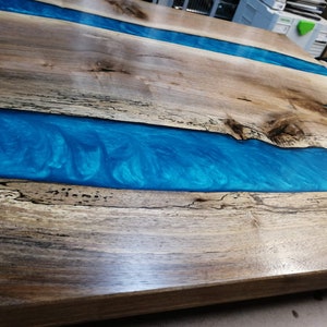 Puede incluir: Una mesa de madera con un r&iacute;o de resina epoxi azul que la atraviesa. La madera tiene una veta natural y la resina tiene un patr&oacute;n de remolino.