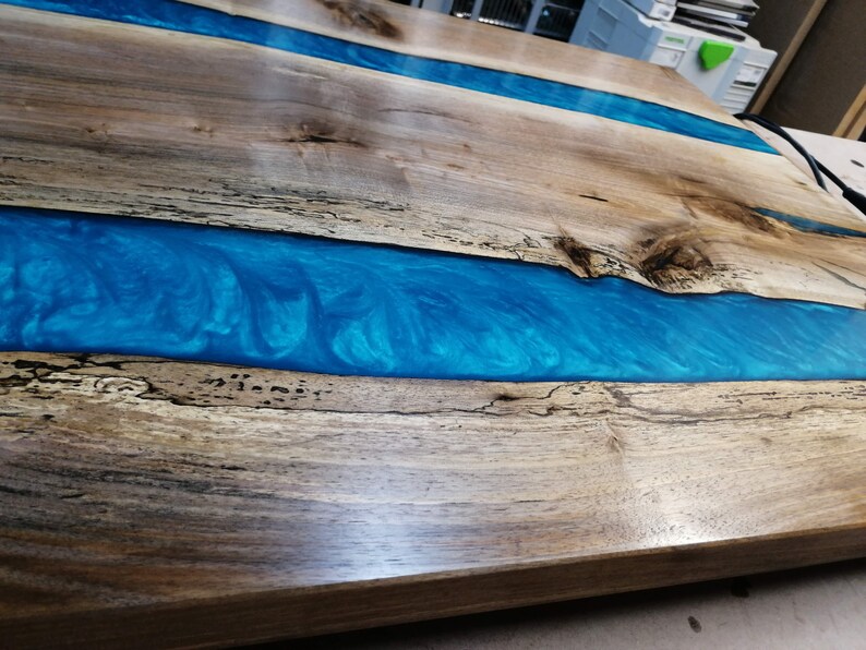 Puede incluir: Una mesa de madera con un r&iacute;o de resina epoxi azul que la atraviesa. La madera tiene una veta natural y la resina tiene un patr&oacute;n de remolino.