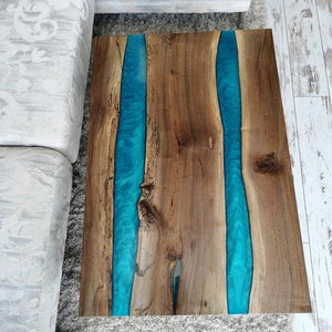 Puede incluir: Una mesa de centro de madera con un acabado de madera natural y dos incrustaciones de resina azul. La mesa tiene una forma rectangular y est&aacute; sobre una alfombra blanca.