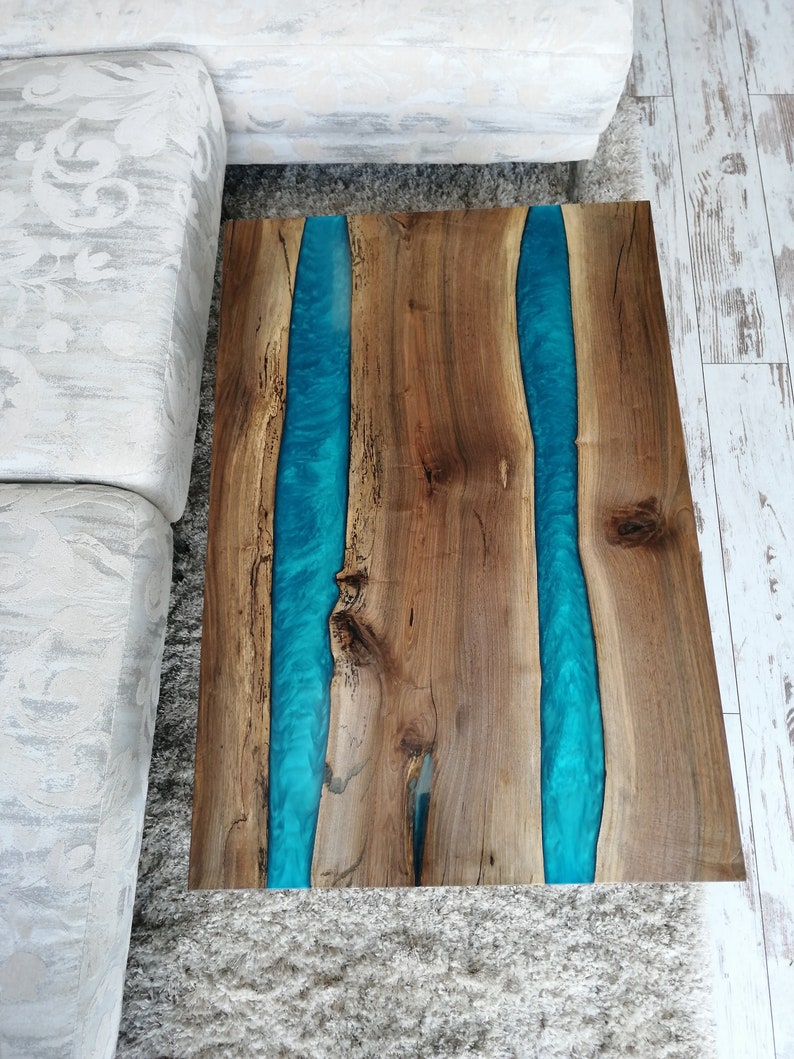 Puede incluir: Una mesa de centro de madera con un acabado de madera natural y dos incrustaciones de resina azul. La mesa tiene una forma rectangular y est&aacute; sobre una alfombra blanca.