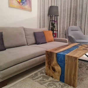 May include: A wooden coffee table with a blue epoxy resin river running through the center. The table has a black metal frame and is sitting on a gray rug in a living room.