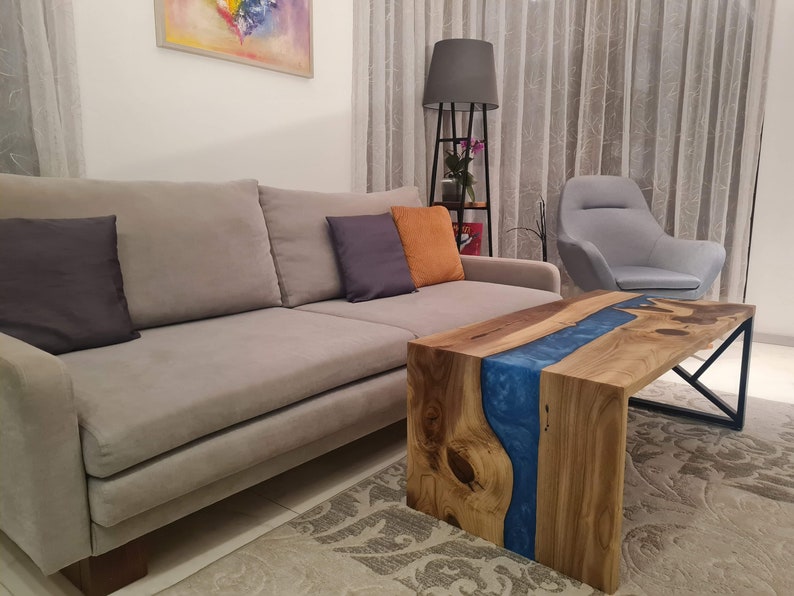 May include: A wooden coffee table with a blue epoxy resin river running through the center. The table has a black metal frame and is sitting on a gray rug in a living room.